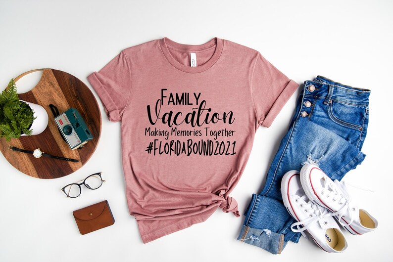 May include: A dusty rose t-shirt with the text "FAMILY Vacation Making Memories Together #FLORIDABOUND2021" printed in black. The shirt is laid flat on a white surface with a pair of blue jeans, white trainers, a wooden chopping board, a camera, a watch, and glasses.