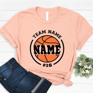 May include: A peach-colored t-shirt with a black and orange basketball graphic. The graphic includes the text "TEAM NAME" and "NAME" with the number "#16" below the basketball.