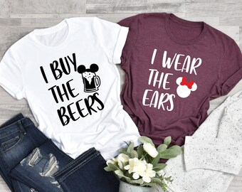 May include: Two white t-shirts with black text and graphics. The first shirt says "I buy the beers" with a cartoon beer mug and Mickey Mouse ears. The second shirt says "I wear the ears" with a cartoon Minnie Mouse head.
