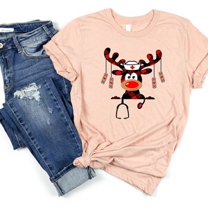 May include: A light pink t-shirt featuring a cartoon reindeer with a nurse's hat, red nose, and plaid antlers. The reindeer is holding a stethoscope. The shirt is paired with blue denim jeans.
