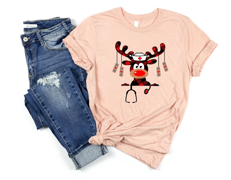 May include: A light pink t-shirt featuring a cartoon reindeer with a nurse's hat, red nose, and plaid antlers. The reindeer is holding a stethoscope. The shirt is paired with blue denim jeans.