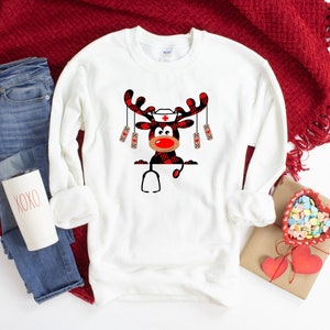 May include: White sweatshirt featuring a cartoon reindeer design. The reindeer has a red nose, a nurse's cap, and a stethoscope. The antlers and parts of the reindeer are in a red and black plaid pattern. The sweatshirt is paired with blue jeans and a red blanket.