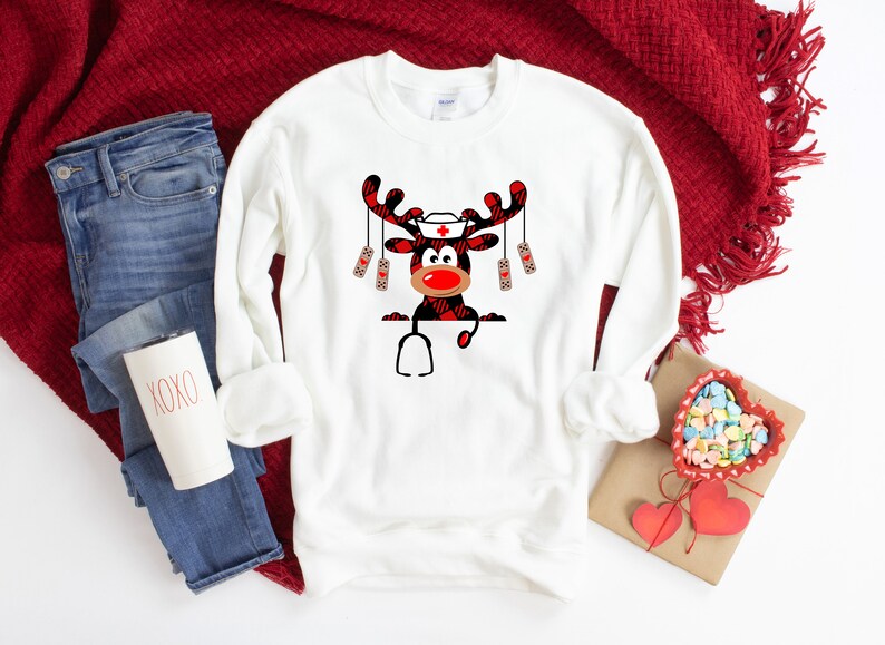 May include: White sweatshirt featuring a cartoon reindeer design. The reindeer has a red nose, a nurse's cap, and a stethoscope. The antlers and parts of the reindeer are in a red and black plaid pattern. The sweatshirt is paired with blue jeans and a red blanket.