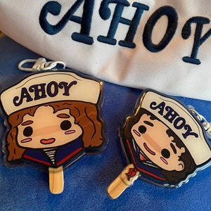 May include: Two cartoon character keychains shaped like popsicles, featuring a sailor hat with the word "AHOY". The keychains have a wooden stick and a white metal clasp. A white sailor hat with the word "AHOY" embroidered in navy blue is in the background.