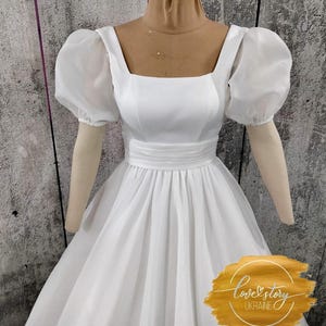 May include: A white wedding dress with a square neckline and puffed sleeves. The dress has a fitted bodice and a full skirt. The dress is on a mannequin and the background is a textured wall.