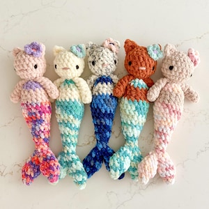 May include: Five handmade crochet mermaid dolls with cat-like ears and small flowers on their heads. Each doll features a soft yarn body with a distinct color scheme, including pink, cream, grey, brown, and beige, paired with textured tails.