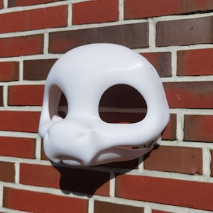 Könnte beinhalten: Weiße leere Felltiermaske mit großen Augenlöchern und einer Schnauze. Die Maske besteht aus einem glatten, harten Material und ist bereit zum Bemalen.