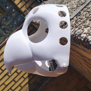 May include: White plastic mask with multiple holes and a spring attached to the side. The mask is shaped like a bird's head.
