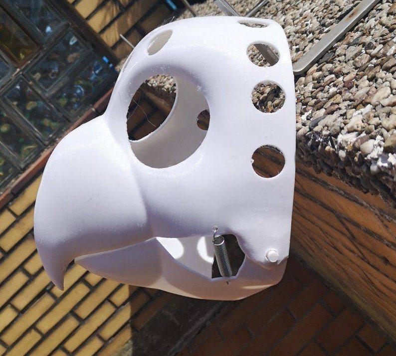May include: White plastic mask with multiple holes and a spring attached to the side. The mask is shaped like a bird's head.
