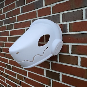 Puede incluir: Máscara de plástico blanco con forma redondeada y superficie texturizada. La máscara tiene una gran abertura para los ojos y una abertura más pequeña para la boca. La máscara está unida a una pared de ladrillo.