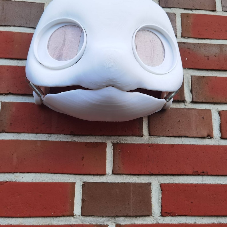 May include: White plaster mask with large round eyes and a small mouth. The mask is attached to a brick wall.