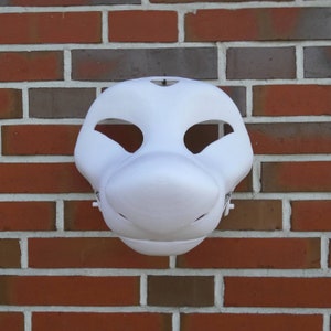 May include: A plain white fursuit mask with large eye openings and a pronounced snout. The mask is mounted on a red brick wall, highlighting its smooth surface and potential for customisation. The mask has small holes for the nose and mouth.