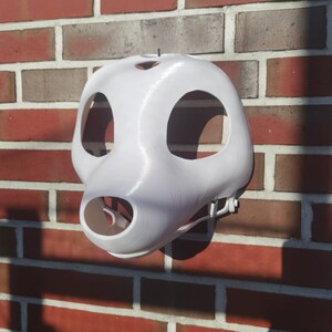 May include: White 3D printed animal mask with holes for eyes and nose. The mask is hanging on a brick wall.