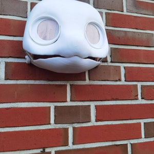 May include: White plaster mask with two large eye holes and a small mouth opening. The mask is hanging on a brick wall.