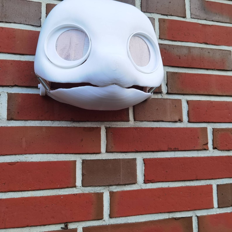 May include: White plaster mask with two large eye holes and a small mouth opening. The mask is hanging on a brick wall.
