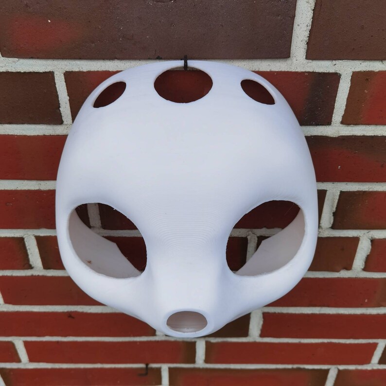 May include: White plastic mask with multiple holes and a rounded shape. The mask is hanging from a hook on a brick wall.