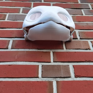 May include: White plastic mask with two round eye holes and a small mouth opening. The mask is attached to a brick wall.
