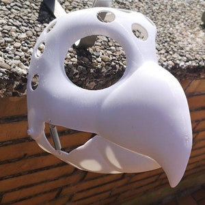 May include: White plastic mask with a beak and holes around the eyes. The mask is hanging from a hook.