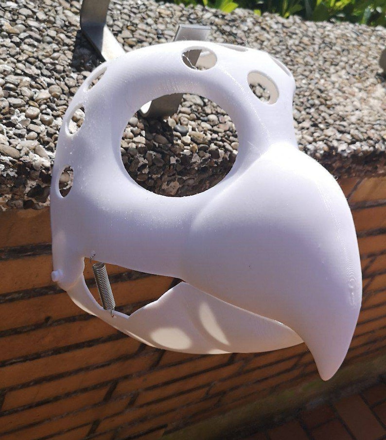 May include: White plastic mask with a beak and holes around the eyes. The mask is hanging from a hook.