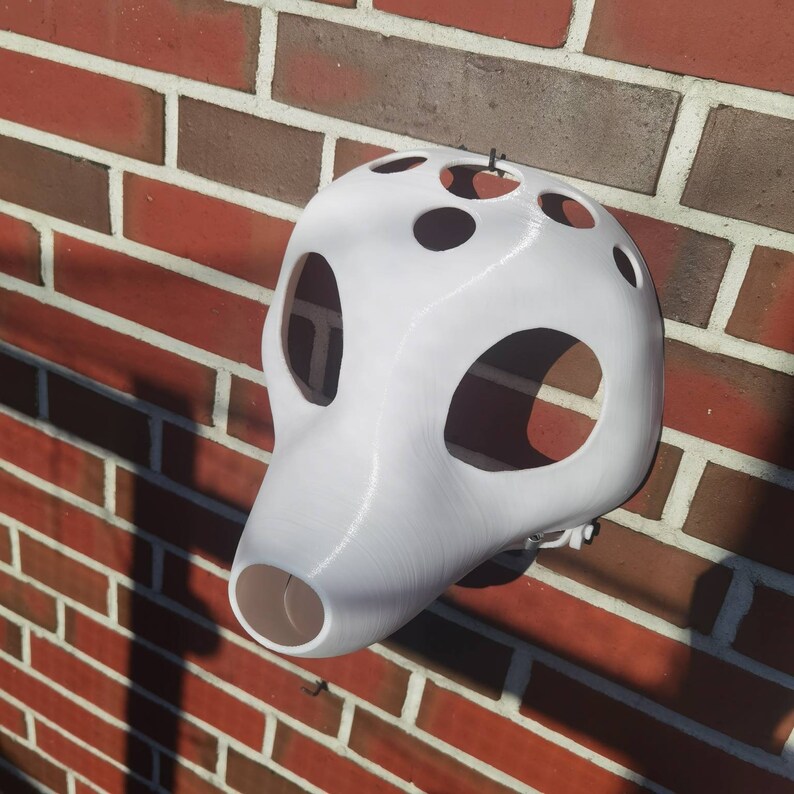 May include: White plastic mask with multiple holes and a rounded shape. The mask is hanging on a brick wall.
