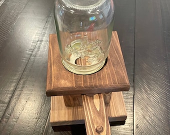 May include: A clear glass jar sits on a stained wooden stand. The stand is constructed from rectangular pieces of wood, joined together to form a sturdy base. The jar has a decorative design on the bottom. The wood has a rich, dark brown color.