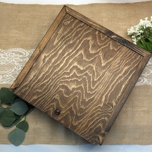 Può includere: Una scatola di legno marrone scuro con un bordo leggermente rialzato attorno al coperchio. La scatola presenta una venatura del legno naturale e una piccola manopola di metallo sul davanti.