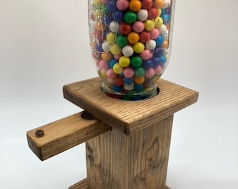 May include: A wooden gumball dispenser with a clear glass jar filled with colorful gumballs. The dispenser has a wooden lever that dispenses the gumballs.