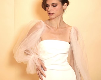 Puede incluir: Un vestido de novia blanco sin tirantes con un corpiño ajustado y una falda fluida. El vestido presenta mangas transparentes y abullonadas con delicados volantes en los puños.