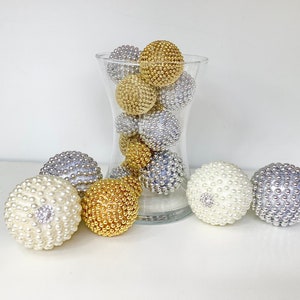 K&ouml;nnte beinhalten: Eine klare Glasvase gef&uuml;llt mit dekorativen Kugeln in Gold, Silber und Wei&szlig;. Die Kugeln sind aus einem strukturierten Material, das an Perlen erinnert.