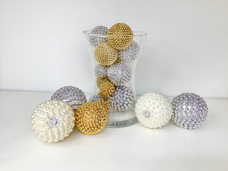 K&ouml;nnte beinhalten: Eine klare Glasvase gef&uuml;llt mit dekorativen Kugeln in Gold, Silber und Wei&szlig;. Die Kugeln sind aus einem strukturierten Material, das an Perlen erinnert.