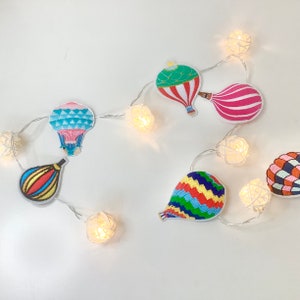 Puede incluir: Una cadena de luces blancas con pequeñas bombillas redondas y tejidas. Cada bombilla está decorada con un parche bordado de globo aerostático de colores. Los globos son de varios colores y patrones, incluyendo rojo, rosa, azul, verde, amarillo y blanco.