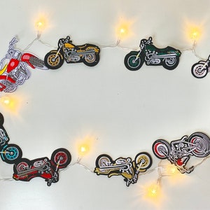 Puede incluir: Ocho parches de motocicleta coloridos con diferentes diseños están enhebrados en una cadena de luces blanca. Los parches son de varios colores, incluyendo rojo, azul, verde, amarillo y negro.