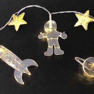 May include: A string of lights with clear plastic astronaut, rocket, star, and planet shapes. The lights are warm white and the shapes are illuminated.