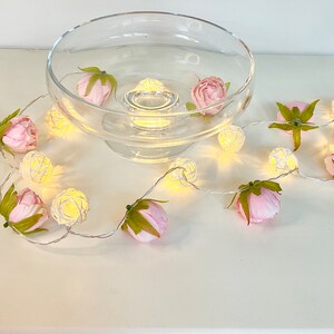 May include: A string of white fairy lights with small, round, woven wire bulbs. The lights are wrapped around a clear glass bowl and pink artificial flowers.