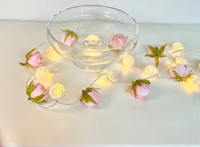 May include: A string of white fairy lights with small, round, woven wire bulbs. The lights are wrapped around a clear glass bowl and pink artificial flowers.