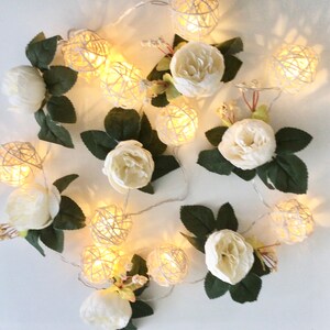 May include: A string of white fairy lights with white paper flowers and woven ball lights. The lights are strung together with a clear wire.
