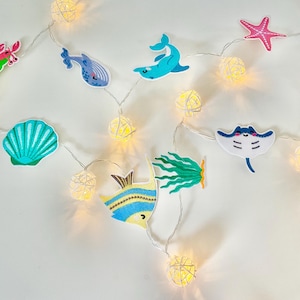 May include: A string of warm white fairy lights with sea creature decorations. The decorations include a pink starfish, a blue dolphin, a purple jellyfish, and a blue and yellow fish. The lights are on a white background.