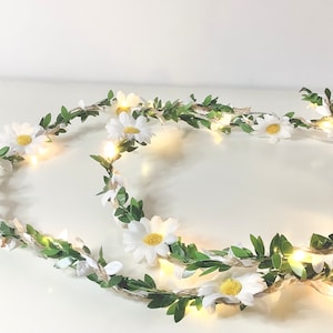 May include: A string of white daisy lights with green leaves, wrapped around a white surface. The lights are strung on a brown twine.