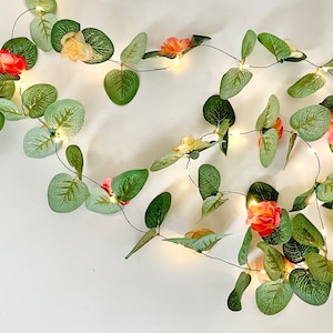 Puede incluir: Una cadena de luces con hojas verdes y flores rosas y amarillas. Las luces están enhebradas en un cable y son perfectas para agregar un toque de fantasía a cualquier espacio.