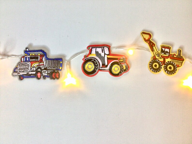 May include: A string of white fairy lights with yellow bulbs, decorated with felt patches of a red tractor, a blue dumper truck, and a yellow excavator.