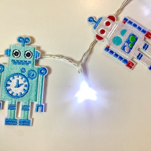 May include: A string of white fairy lights with blue and white robot-shaped ornaments. The robots have embroidered details and are lit by the white lights.