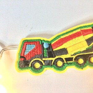 May include: A yellow, green, red and black embroidered patch of a cement mixer truck. The truck has a rotating drum and four wheels.