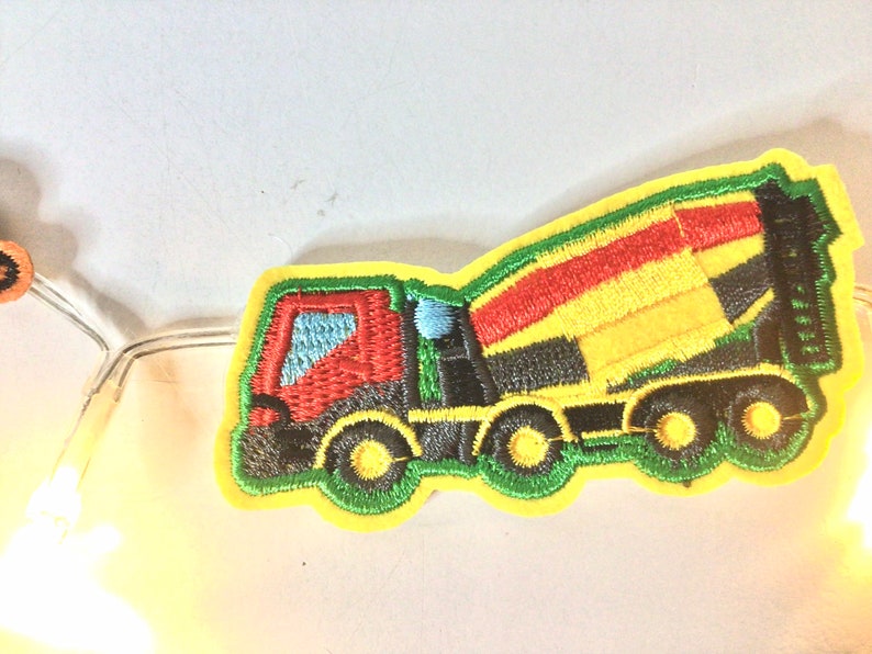 May include: A yellow, green, red and black embroidered patch of a cement mixer truck. The truck has a rotating drum and four wheels.
