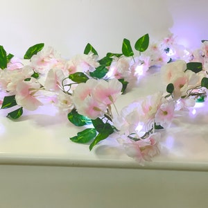 Puede incluir: Una guirnalda floral con delicadas flores rosas y blancas y hojas verdes. La guirnalda está iluminada con pequeñas luces blancas brillantes, creando un brillo suave. Las flores están agrupadas, creando una apariencia completa y decorativa.