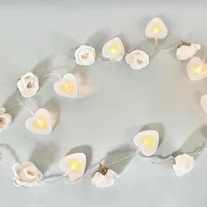 May include: A string of decorative lights featuring alternating white fabric roses and white heart-shaped plastic casings. Each heart contains a small, warm yellow LED light that glows when illuminated. The components are connected by a thin, flexible wire.