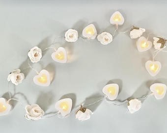 May include: A string of decorative lights featuring alternating white fabric roses and white heart-shaped plastic casings. Each heart contains a small, warm yellow LED light that glows when illuminated. The components are connected by a thin, flexible wire.