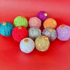 Könnte beinhalten: Eine Sammlung farbenfroher Garnkugel-Ornamente in den Farbtönen Türkis, Grün, Rot, Rosa, Silber, Orange, Lila, Weiß und Gold. Jedes Ornament hat eine goldfarbene Kappe. Die Ornamente sind auf rotem Hintergrund angeordnet.