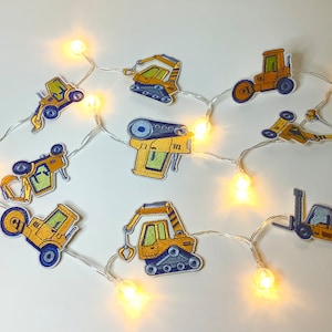 Könnte beinhalten: Eine Lichterkette mit weißen Lichtern und gelben Glühbirnen, verziert mit gelben und blauen Aufklebern von Baufahrzeugen. Die Aufkleber zeigen einen Bulldozer, einen Bagger, einen Radlader, einen Kipplaster und einen Gabelstapler.