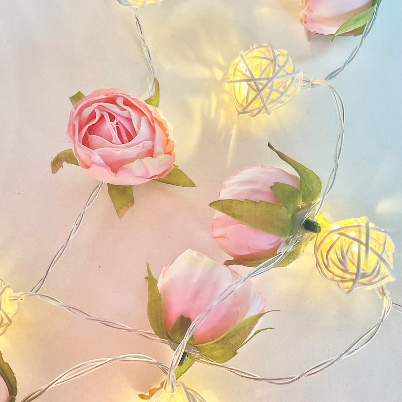 May include: A string of fairy lights with pink fabric rose flowers and woven ball lights. The lights are strung on a white wire.
