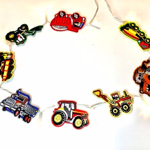 May include: A string of white fairy lights with colourful felt construction vehicle cut-outs. The vehicles include a dumper truck, a tractor, a bulldozer, a cement mixer, and a digger.